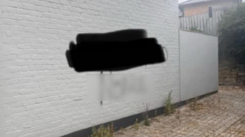 Graffiti sprayed on a white wall of a housing building. There are words which have been blurred out with black and a clear blurring. There is a small brick yard in front of the side of the building. 