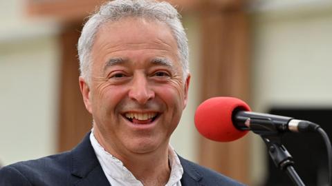 A smiling Frank Cottrell-Boyce, with short grey hair and wearing a dark jacket and white shirt, stands in front of a red microphone