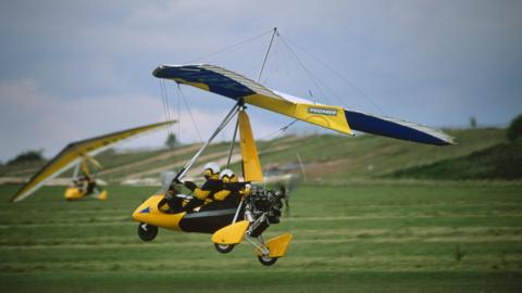 A Pegasus Quantum 15 light aircraft. The black and yellow aircraft has triangular wings above a trike cockpit. A propeller is at the rear of the vehicle. Two people are sitting in the cockpit.