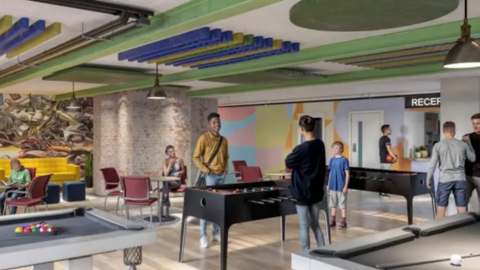 A picture of one of the planned rooms within the youth centre. There is a number of people stood around tables, including football and pool tables. There is also a small seating area to the left of the room.