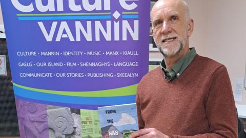 David Callin, who has grey hair and a grey beard and it wearing a red jumper. He is standing next to a banner that reads Culture Vannin. 