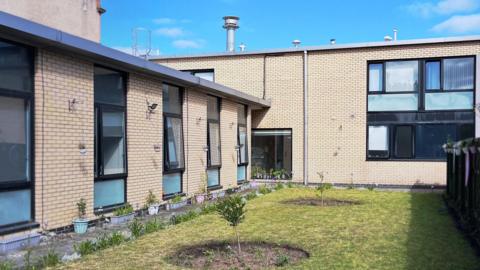 Accessible courtyard garden between brick residential buildings, featuring a well-kept lawn with newly planted small trees, flower pots lining the building walls, and hanging planters on a dark fence under a bright blue sky with scattered clouds.