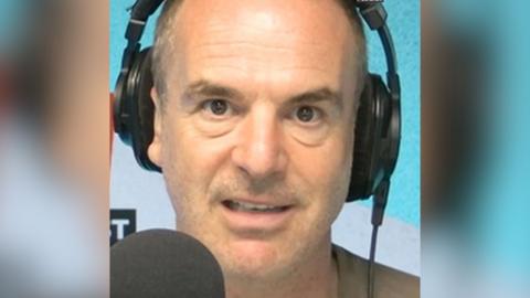 A man with short hair looks into the camera as he talks into a microphone