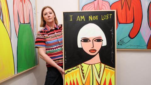 Margo McDaid dressed in a colourful shirt, stood in front of a wall with two of her paintings. She is holding another large painting of a woman in yellow clothing, which has the words 'I am not lost' on it.