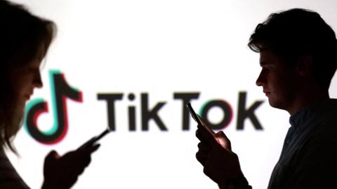 Teenagers pose for a photo while holding smartphones in front of a TikTok logo
