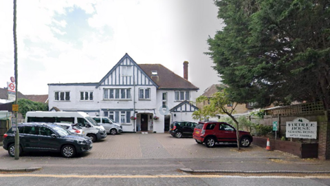 Street view image of a large white care home. There are six vehicles parked in the car par in front of the building and trees to the right