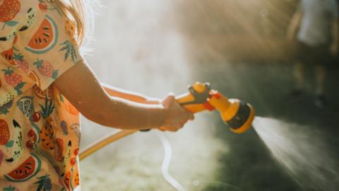 A girl wearing a patterned top holds a yellow garden hose with an attachment. She sprays water from the hose attachment.  A man stands in the top right background of the picture.