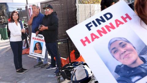 Three volunteers putting up flyers of missing Maui woman Hannah Kobayashi in Los Angeles stand near a wall, with one holding a poster that reads "Find Hannah". A more close-up poster is featured on the right side of the picture, featuring a photo of Ms Kobayashi smiling.
