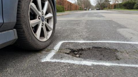 A pothole in a quiet road marked in white paint next to a car wheel. The pothole is an uneven shape and is sunken into a road which is elsewhere patched up in different areas.