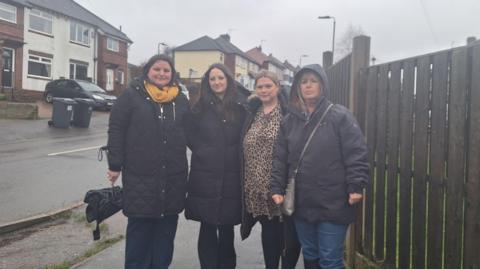 Four women stood close together in a row. They are wearing jackets, with one woman who has her hood up. They are stood on the pavement of a road with a fence and houses in the background. 