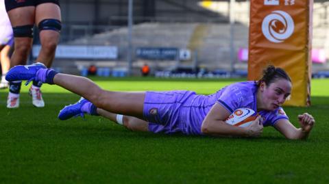 Nel Metcalfe lying on the grass over the tryline with the ball tucked under one arm and her fist clenched in the other after scoring a try