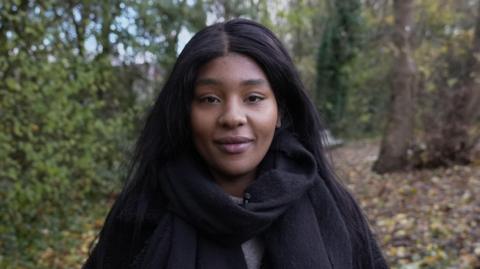 A young black woman looks directly into the camera. She is standing in a forest with the trees and leaves on the ground behind her slightly blurred. She has long black soft and straight hair, parted in the middle. She is wearing a large black scarf and there is a hint of a grey jumper underneath. She has a slight smile on her face.