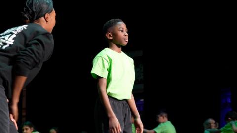 A young boy stands on a stage wearing a green T-shirt and black trousers.