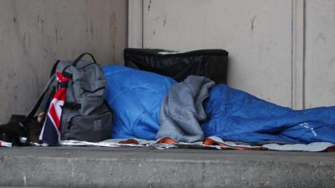 Rough sleeper in blue sleeping bag next to a rucksack on hard concrete floor