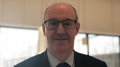 Neil Guckian is wearing a pink and white striped tie and wearing black-rimmed glasses. He is wearing a black jacket and is sitting in a purple and brown chair in a room with a number of people. 