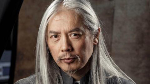 Clem So with white long hair and brown eyes with a small goatee beard. He is wearing a grey suit.