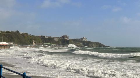 Choppy waves crashing on the shore from right to left with a sea wall and cliff edge in background under a blue sky