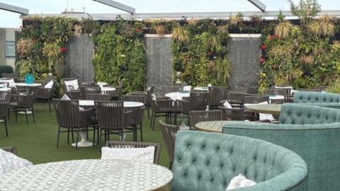 Roof area of a building being used as a restaurant. In the foreground is a turquoise-coloured high-backed chair in a crescent shape, with a table. Behind are a number of tables and chairs. There is a green carpet. The sky is visible.