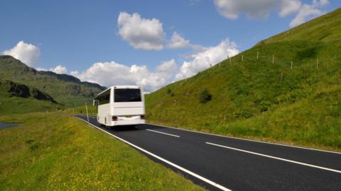 A white coach drives along a road with grassy verges on each side and a mountain in the background. 