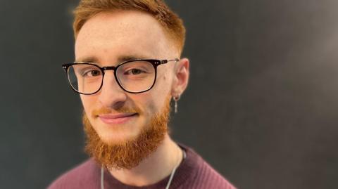 Ginger haired man with a beard and a half-smile, wearing a burgundy ribbed jumper and metal neck chain and large round glasses sitting in front of a black background.