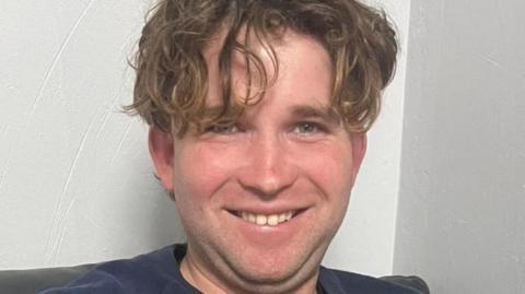 James Nunan has long, curly brown hair drooping downwards, partially covering one eye. He is smiling in front of a white wall and wearing a navy T-shirt.