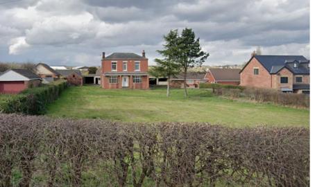 A rural or semi-rural residential scene with a large open grassy field in the foreground. The field is unevenly cut and bordered at the front by a low hedge made of thin, leafless branches, forming a natural boundary along the roadside or edge of the property.
In the middle of the image, there is a two-storey red brick house positioned centrally within the field. The house has a symmetrical front with white-framed windows and a central front door. The roof is dark grey and slopes gently, with chimneys visible on either side. Behind and beside the main house, there are additional outbuildings and structures, including what appears to be a barn or garage with a wide opening.