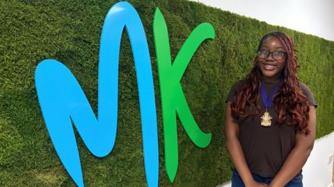 A young woman wearing glasses and a ceremonial badge stands with folded hands beside a large blue-and-green logo attached to a textured green wall, with office space visible in the background.