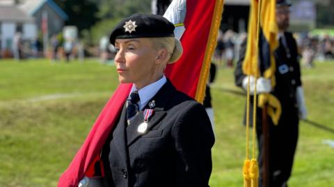 McCabe in uniform carrying the Manx flag. There is green grass around and other flag bearer standing to attention.