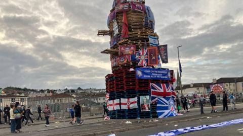 A large unlit bonfire covered in British and Northern Ireland flags beside a road. There are people milling around and cream-coloured blocks of flats in the background.