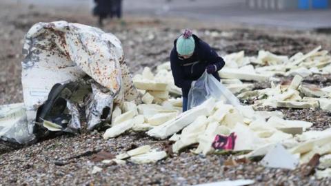 A person putting white blocks of foam into a large, clear plastic bag on a pebble beach