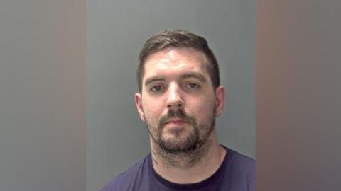 Jacob Hall is looking at the camera in a police custody shot. he has short dark brown hair, brown eyes and a beard and moustache. He is wearing a dark blue T-shirt