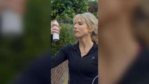 Lucy Punch as Amanda is wearing a black gym top and spraying a bottle of makeup setting spray on her face.