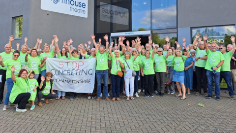 A group of people from the Staunch campaign all wearing matching green tops while standing with their arms raised in the air. They are outside the Lighthouse Theatre. Some of the people are holding a white banner which reads - Stop Concreting Northamptonshire.