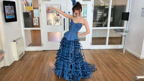 Tabitha shown posing in the dress she designed at school. It is a denim blue, with a long structured corset and a layered skirt. 