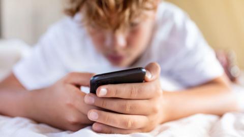 Teenage boy holding a phone lying on a bed. Phone is close to camera and the boy's head is blurred.