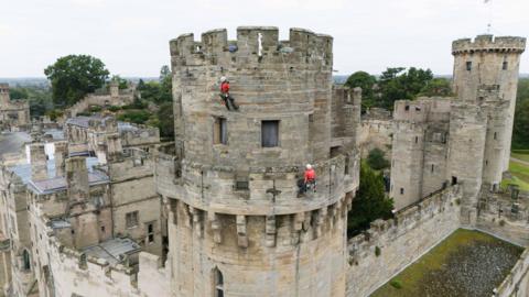 Two people in red t-shirts and white helmets are abseiling stone walls at Warwick Castle