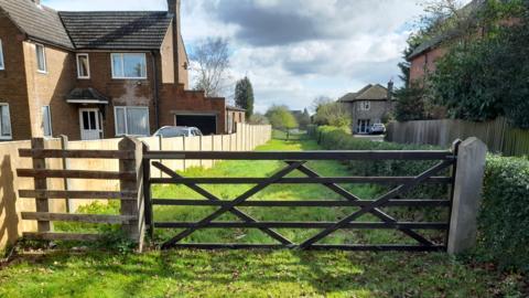 Access to former cathedral quarry site, Riseholme Road, Lincoln. The picture shows a wooden gate across a narrow stretch of grass, with houses either side.