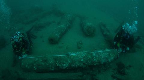 Underwater image of two divers measuring a barnacle-covered canon on the sea bed.