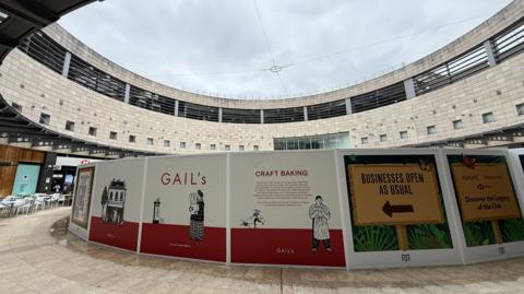 Hoarding for bakery chain Gail's erected in a circular courtyard. The hoarding has colourful branding on it, coloured white and red. It is cloudy above.