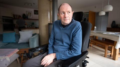 A man sat at his home in a wheelchair wearing a blue jumper.