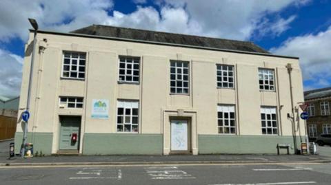 The Outside In building. It is the front of a two-storey building that has two doorways, one at the front left and the other central. Half of the first floor is painted sage green, and the rest of the building is painted cream. there are 9 windows along the front of the building and a sloped roof. There is a sign for the nursery between two windows, at the left handside of the building.