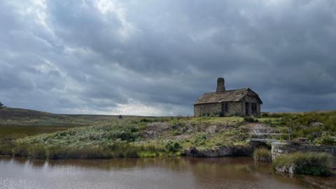 A small stone building on grassland behind a small body of water and beneath a dark, cloudy sky