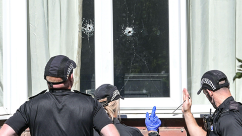 Three police officers in black uniforms and baseball style caps carry out a forensic examination. They are looking at two bullet holes in the window of a house 