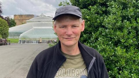 Allan Richardson - man in his 60s - with grey hair smiling at the camera. He is wearing a black baseball cap, a green t-shirt and black zip-up fleece. He is standing in front of the glass structure of the Kibble Palace in Glasgow's Botanic Gardens and there are trees and benches in the background.