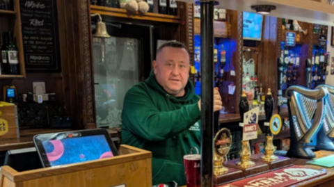 A man in a green hoodie standing behind a bar and pulling a pint. He is looking at the camera.