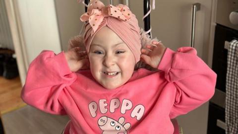 A smiling Amber grinning at the camera. She is wearing a bandanna as her hai grows up, and pink clothes with Peppa Pig on them. She is holding her hands to her ears. 