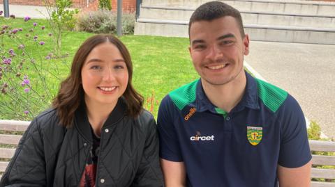 Eimhear O'Neill and Luke MacDaibheid sitting on a bench beside each other. They are both looking at the camera and smiling. Ms O'Neill is wearing a black coat and she has brown hair. Mr MacDaibheid is wearing a green and navy Donegal GAA polo. He has brown hair. 