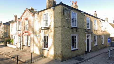 The Fountain is a pub situated on the corner where two roads meet. It is a brick building and has white framed windows and a gold sign on the left that says The Fountain.