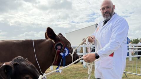 A man wearing a long white coat smiles at the camera. He holds two brown cattle to the left by ropes tied around their heads.