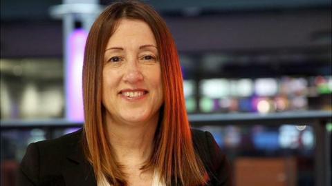 Jane Dodds wearing a black jacket in front of a gallery of TV screens.